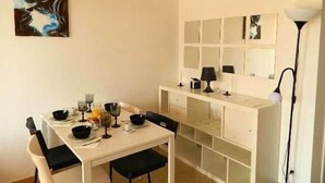 Uma área de refeições com uma mesa branca posta para quatro pessoas, uma cadeira preta, um quadro na parede e um abajur em uma mesinha lateral.