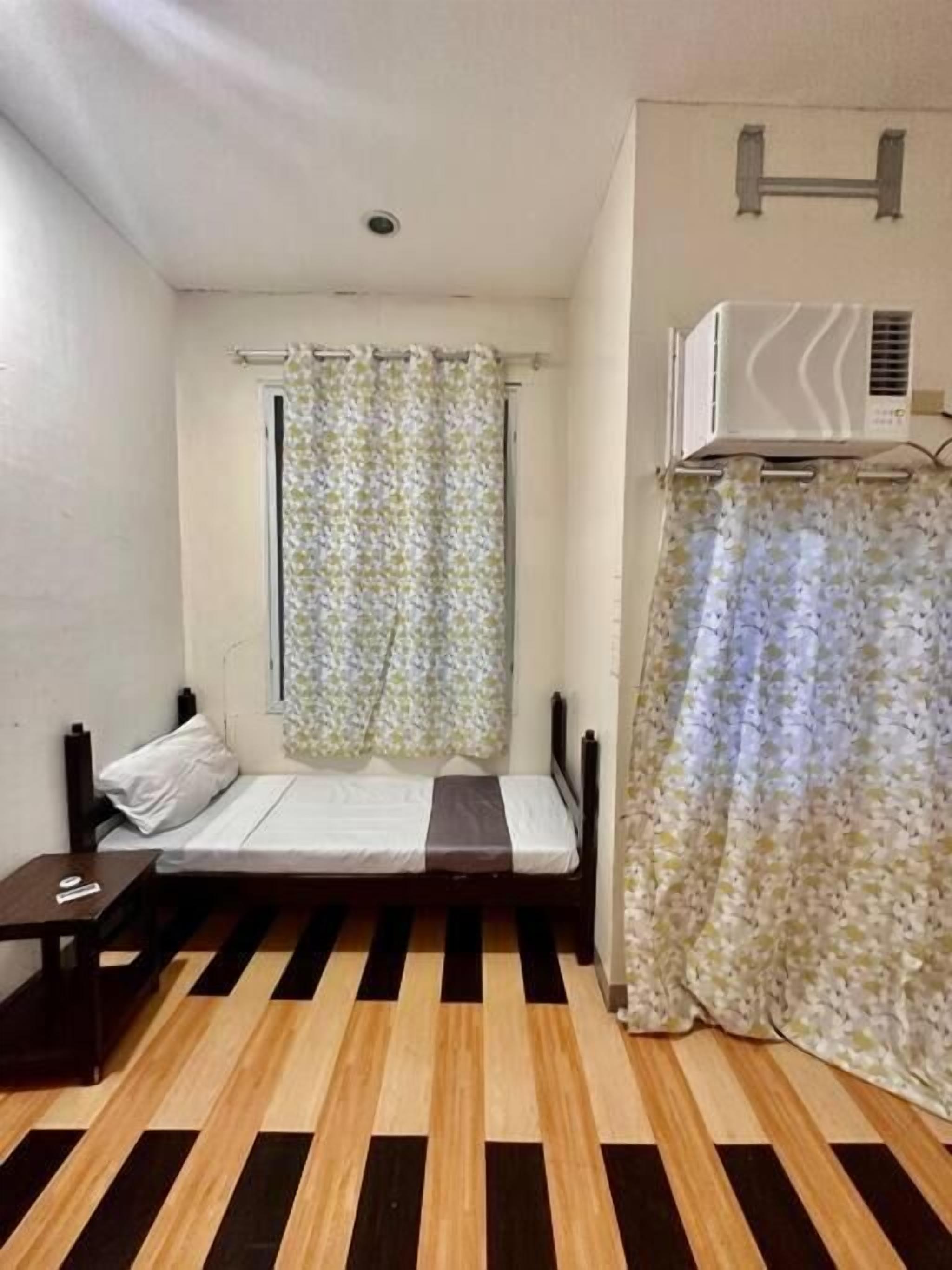 Una habitación pequeña con cama individual, piso de madera, ropa de cama blanca y marrón, una mesita de noche, una ventana con cortina floral y un equipo de aire acondicionado en la pared.