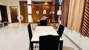 En matplats med ett långt bord och fyra stolar, en fläkt och en soffa i bakgrunden.