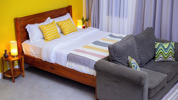 A neatly made bed with a wooden headboard, a grey sofa with patterned cushions, and a bedside table with a lamp.