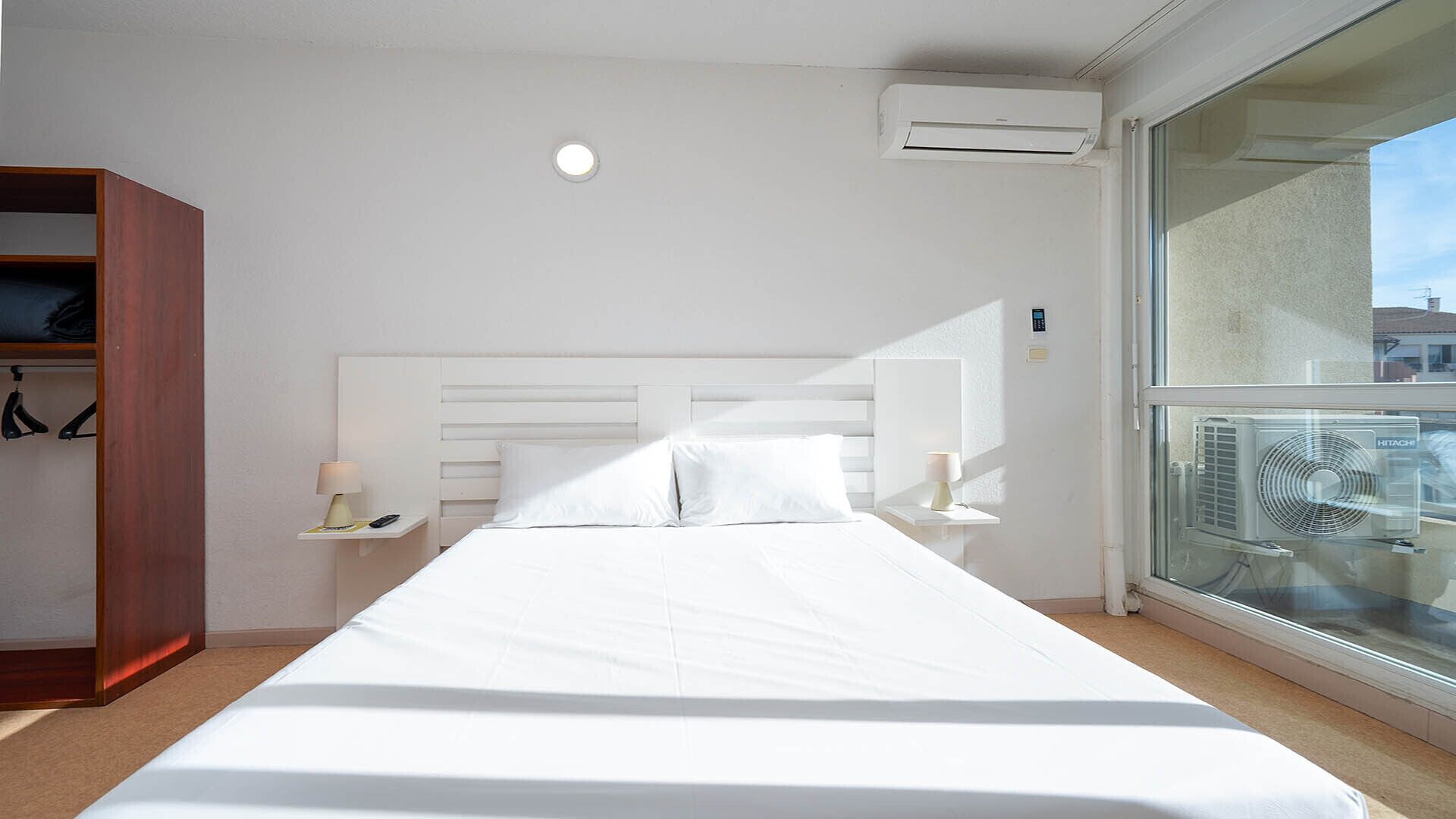 Hotelkamer met een groot bed, een kledingkast, een nachtkastje met een lamp, een airconditioning en een raam met uitzicht op gebouwen.