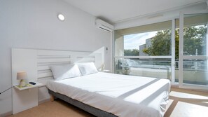 A modern bedroom with a large bed, white bedding, a bedside table with a lamp, and a view of the outdoors through a glass door. - Nice bright studio apartment for 2 people (Agde)