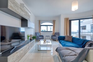 Un salon moderne doté d’un téléviseur à écran plat, d’un canapé, de deux fauteuils, d’une table basse et d’une grande fenêtre avec des rideaux.