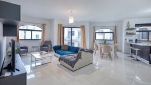Ein modernes Wohnzimmer mit Sofa, Couchtisch und Fernseher. Zwei Fenster mit Vorhängen, eine Küchenzeile mit Mikrowelle sowie ein Esstisch mit Stühlen.