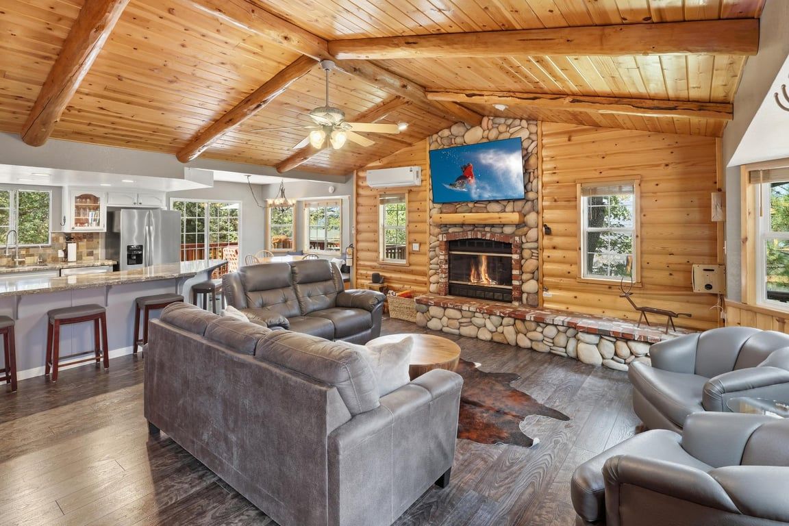 A log cabin interior with a stone fireplace, leather seating, and a kitchen area.