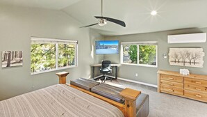 A bedroom with a large bed, a desk, and a chair. There are windows with views of trees and a TV on the wall. - Resort-Style Backyard with Cedar HOT TUB, Sauna, Firepit & Putting Green! PET FRIENDLY! 2 Split Ductless Air Conditioning Units!FREE EV Charger! (Big Bear City)
