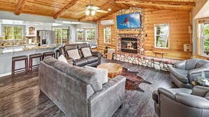 A log cabin interior with a stone fireplace, leather sofas, and a kitchen area. - Resort-Style Backyard with Cedar HOT TUB, Sauna, Firepit & Putting Green! PET FRIENDLY! 2 Split Ductless Air Conditioning Units!FREE EV Charger! (Big Bear City)