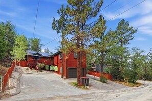 A residential area with a red house, a driveway, and a large pine tree. - Cedar HOT TUB on Front Deck! Resort-Style Backyard w/Sauna, Firepit & Putting Green! PET FRIENDLY! Split Ductless A/C! FREE EV Charger! (Big Bear City)