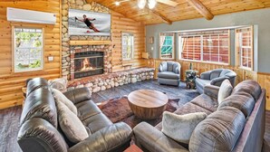 A log cabin interior with a stone fireplace, leather sofas, and a ceiling fan. - Resort-Style Backyard with Cedar HOT TUB, Sauna, Firepit & Putting Green! PET FRIENDLY! 2 Split Ductless Air Conditioning Units!FREE EV Charger! (Big Bear City)