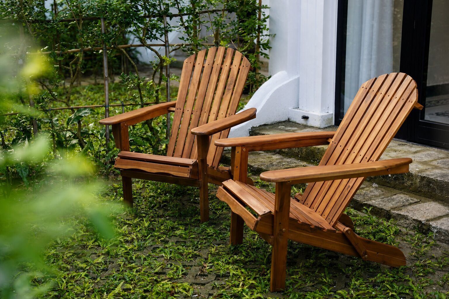 Two wooden Adirondack chairs in a garden setting with a white building in the background.