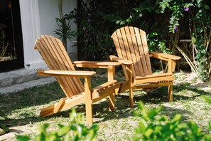 Two wooden Adirondack chairs in a garden setting with a white building in the background. - MUCA Hoian Co-living & Work Retreat (Hoi An)