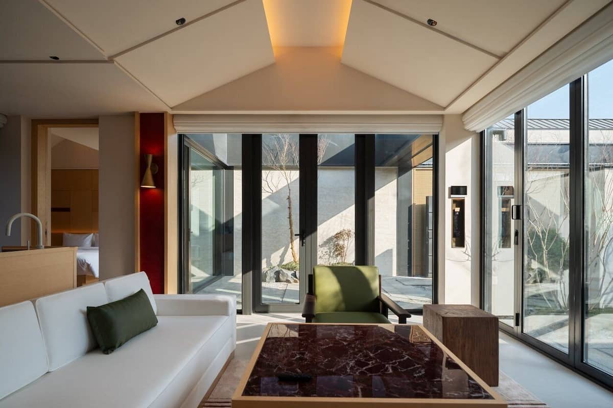 Un intérieur moderne avec un canapé blanc, un fauteuil vert, une table basse en marbre et de grandes fenêtres donnant sur l’extérieur.