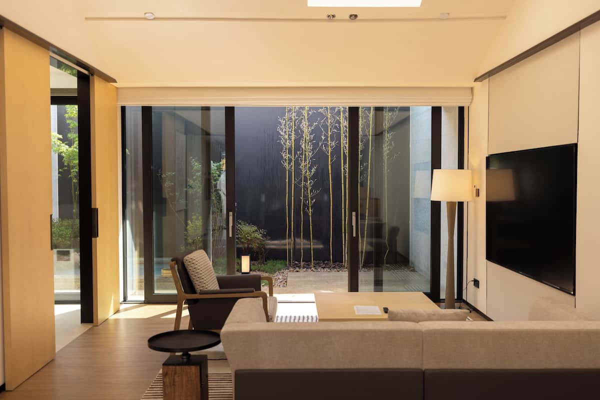 Un salon moderne avec un canapé, un fauteuil, un téléviseur et une vue sur un jardin, visible à travers de grandes portes vitrées.