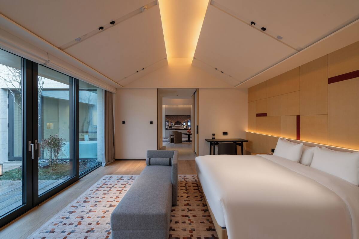 Une chambre d’hôtel moderne dotée d’un grand lit, d’un coin salon, d’un bureau et offrant une vue sur l’extérieur grâce à une porte vitrée.