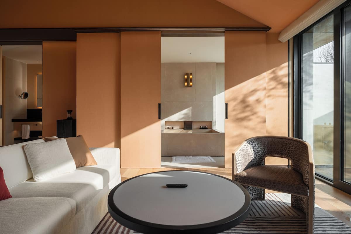 Une chambre d’hôtel moderne avec un canapé, une table basse ronde, un fauteuil à motifs léopard, une grande fenêtre et une vue sur la cuisine.