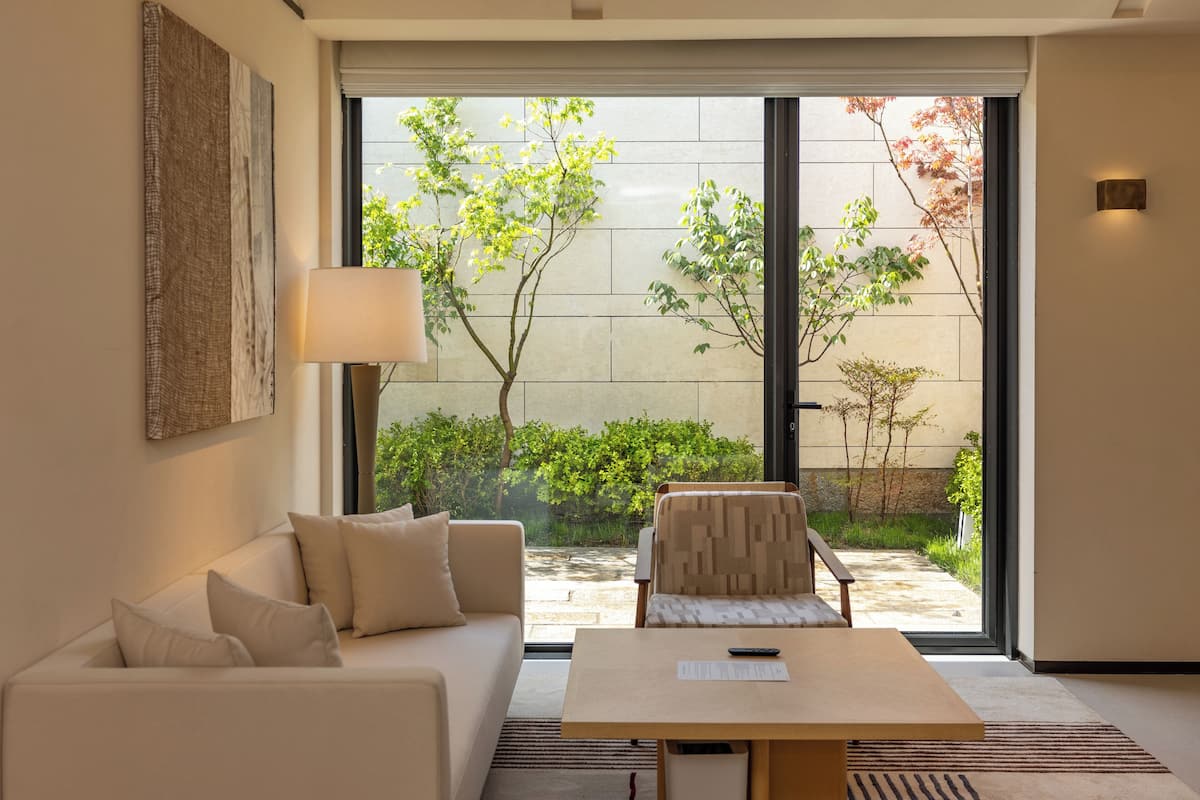 Un salon moderne avec un canapé, une table et une chaise, une grande fenêtre donnant sur un paysage verdoyant et une lampe fixée au mur.