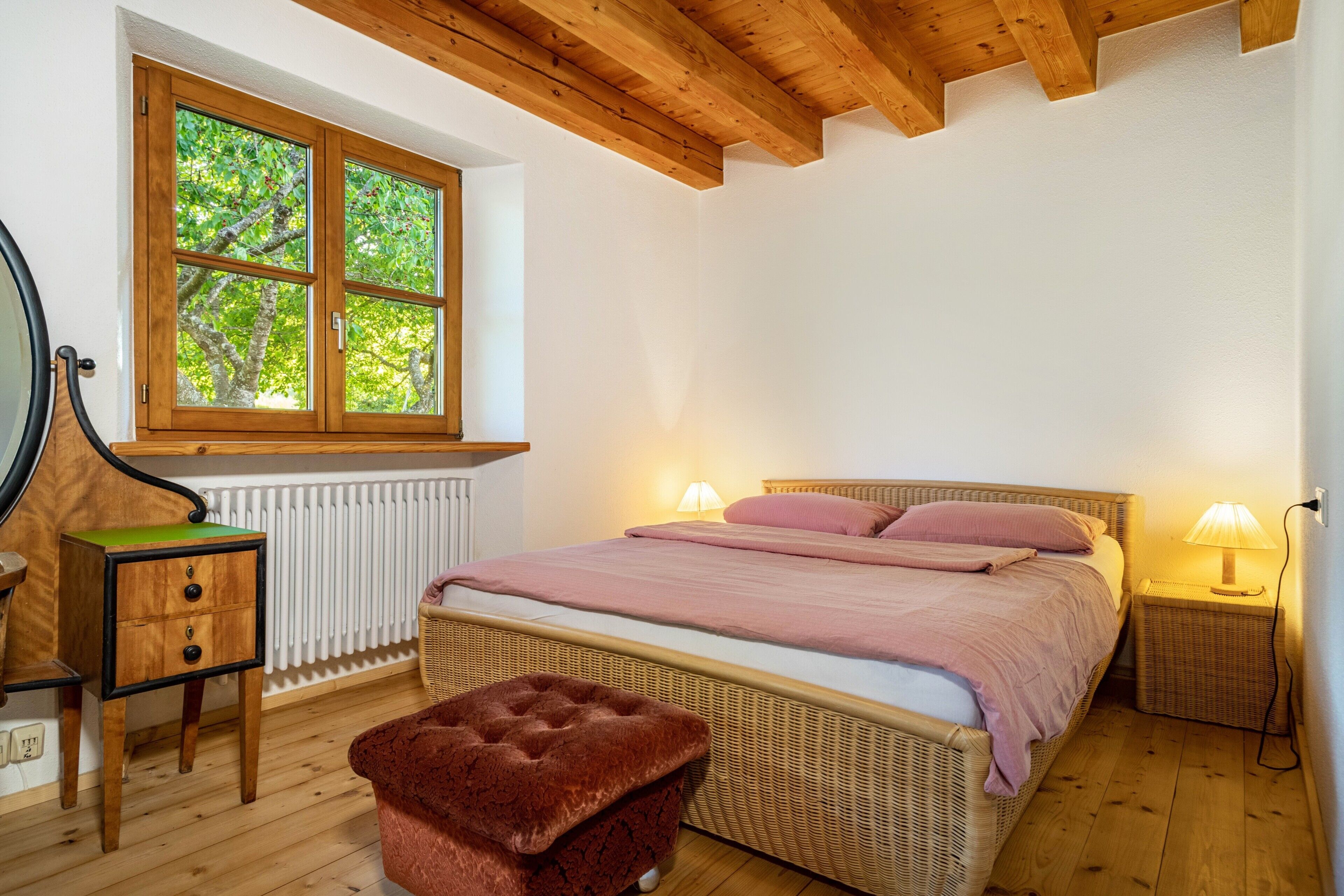 A bedroom with a wooden ceiling, a window with a view of trees, a bed with pink bedding, a nightstand with a lamp, and a small stool.