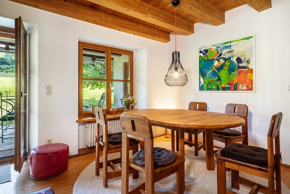 A dining area with a wooden table and chairs, a red ottoman, a hanging light fixture, and a colorful abstract painting on the wall.