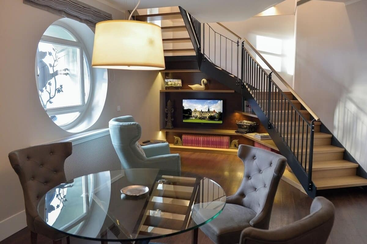 A modern living room with a glass coffee table, two upholstered chairs, a television, and a staircase with a black railing.
