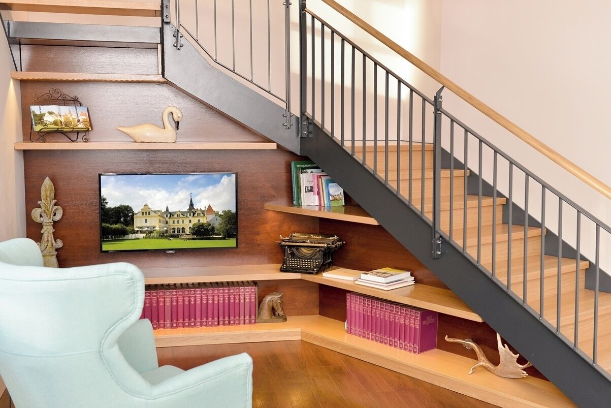 Una escalera con barandilla de madera y balaustres metálicos, flanqueada por estantes con libros, objetos decorativos y un televisor que muestra un paisaje.