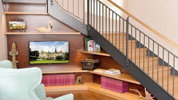 Una escalera con barandilla de madera y balaustres metálicos, flanqueada por estantes con libros, objetos decorativos y un televisor que muestra un paisaje.
