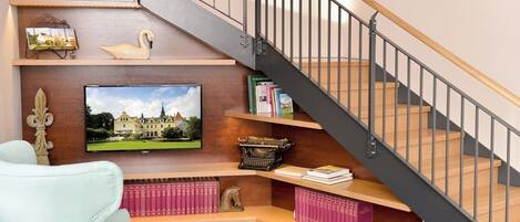 A staircase with a wooden handrail and metal balusters, flanked by shelves with books, decorative items, and a television displaying a landscape.