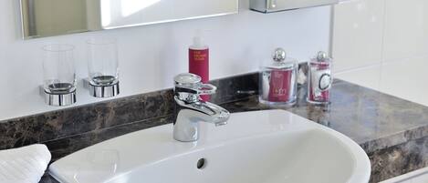 Un baño con lavamanos blanco, grifo cromado y encimera de mármol. Hay dos vasos de vidrio transparente, una botella rosa y una botella roja sobre la encimera.