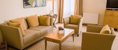 Habitación de hotel con sofá color beige, un sillón más pequeño, una mesa de centro de madera y un televisor sobre un mueble.