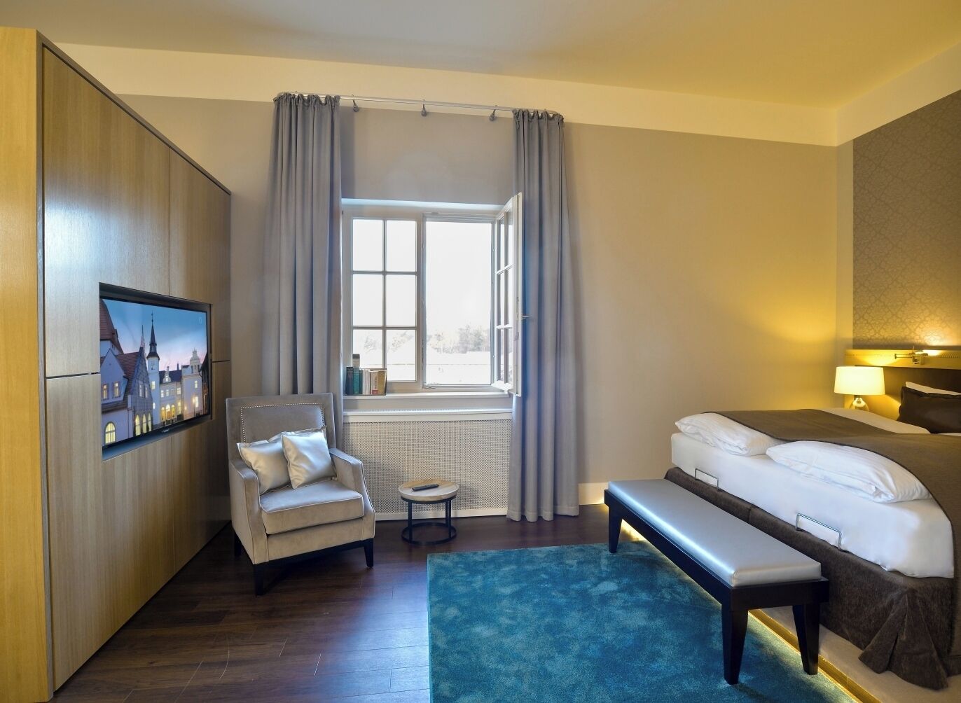Habitació d'hotel amb llit, una cadira, una taula petita, un televisor penjat a la paret i una finestra amb cortines.