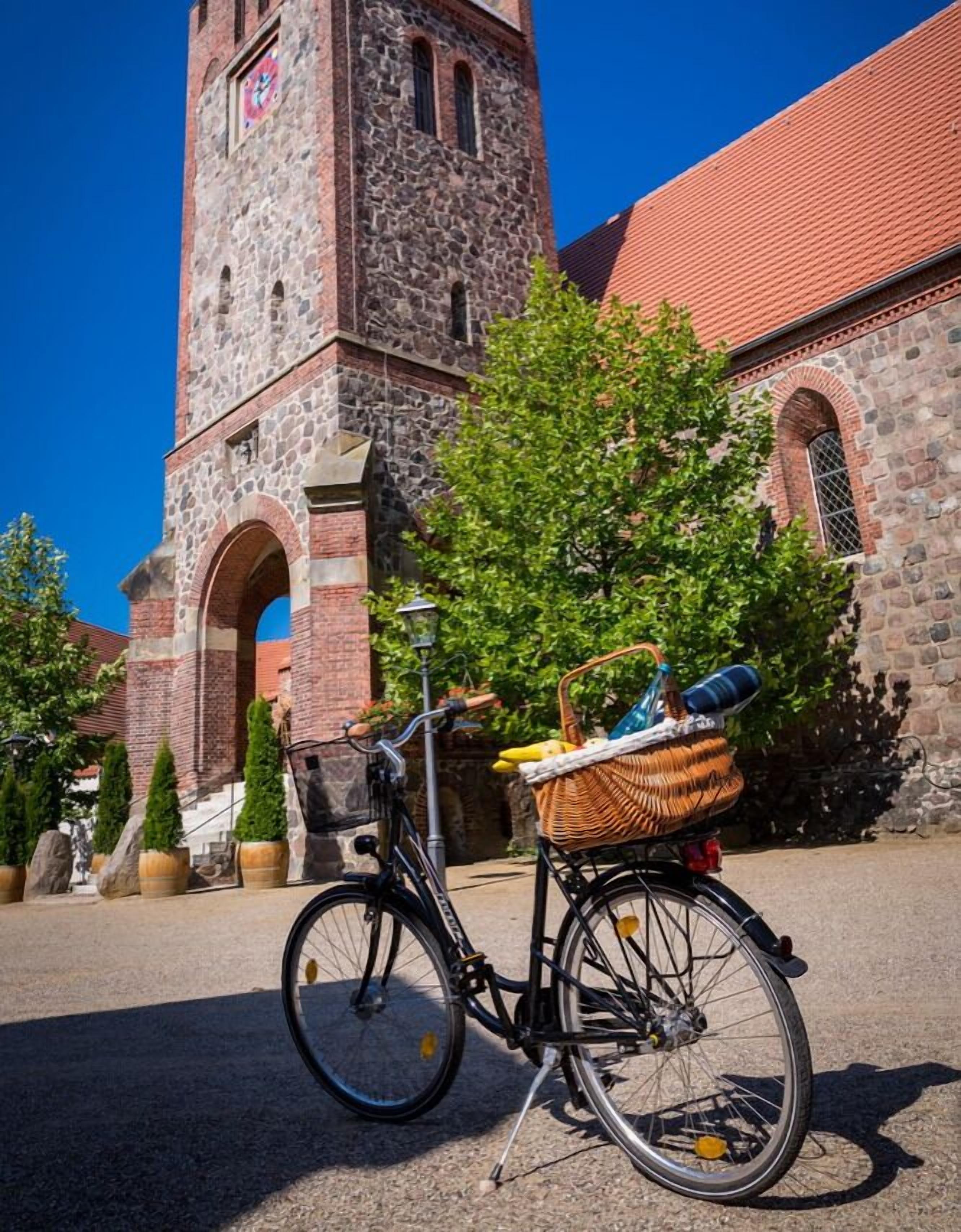 A bicycle with a basket and a wicker basket attached to the back, parked in front of a stone church with a clock tower.