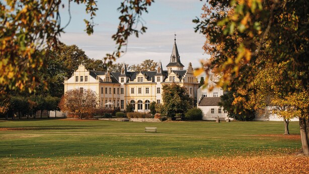 Een groot, historisch gebouw met meerdere torens en een opvallende spits, omgeven door een verzorgd gazon en bomen.