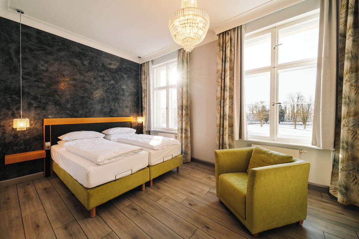 A bedroom with a double bed, a green armchair, a chandelier, and a view of the outdoors through large windows.