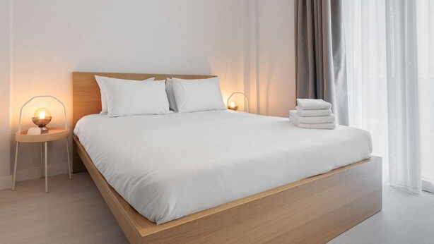 Ein modernes Hotelzimmer mit einem großen Bett, einem hölzernen Kopfteil, weißer Bettwäsche, zwei Nachttischlampen und einem Stapel Handtücher.