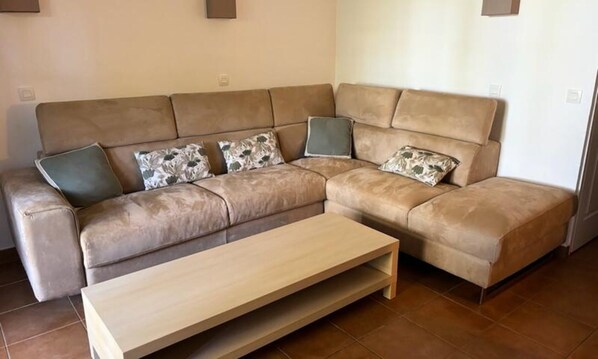 Ein beiges Ecksofa mit floralen Kissen, ein Holztisch und Fliesenboden.