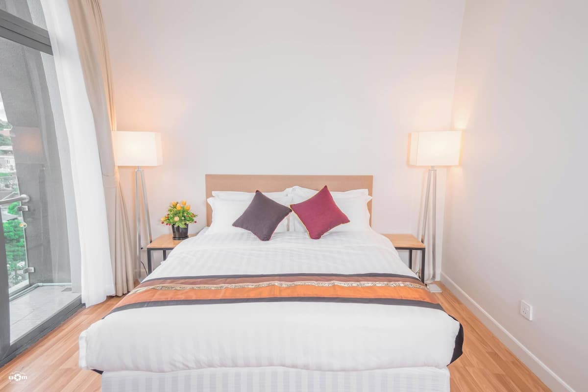 Una cama bien hecha con dos almohadas, una mesita de noche con lámpara y una ventana con cortinas.