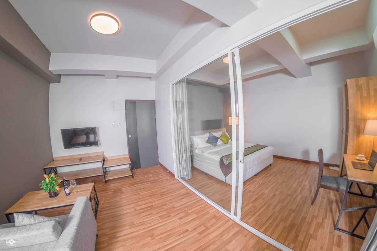 Una habitación de hotel moderna con suelo de madera, un sofá, una mesita con una planta y una botella, una cama con puertas de vidrio correderas, un escritorio con una silla y un televisor en la pared.