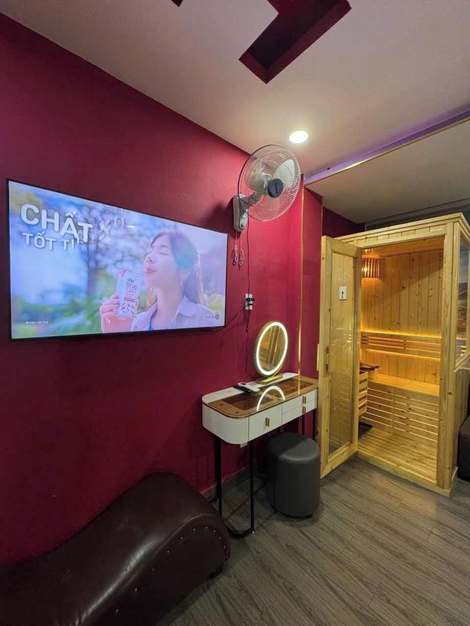 Ein Zimmer mit an der Wand montiertem Fernseher, einem Ventilator, einem Schminktisch mit Spiegel, einer Holztür zur Sauna und einer Lederbank.
