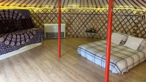 Interior de una yurta tradicional con piso de madera, dos camas con ropa de cama estampada, una mesita y un equipo de aire acondicionado.
