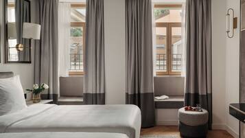 A hotel room with a bed, a bedside table with a lamp, a window with curtains, a small stool with a tray, and a view of the outside.