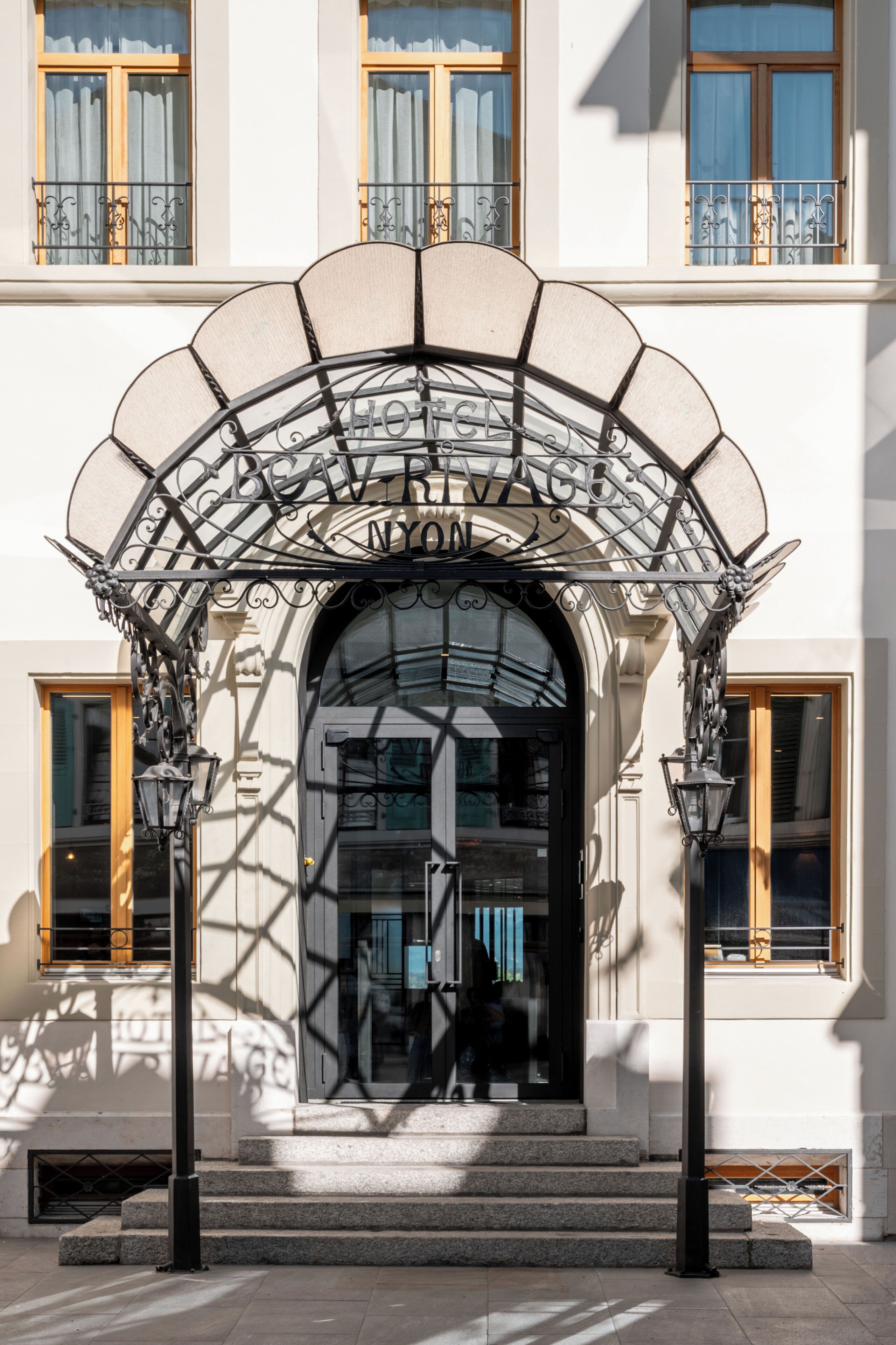 L’entrée d’un hôtel, ornée d’un auvent en fer forgé, avec une enseigne indiquant « HÔTEL DE VOGUE N.Y.O. », et un escalier en pierre menant à la porte.