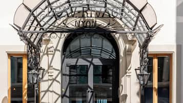 L’entrée d’un hôtel, ornée d’un auvent en fer forgé, avec une enseigne indiquant « HÔTEL DE VOGUE N.Y.O. », et un escalier en pierre menant à la porte.