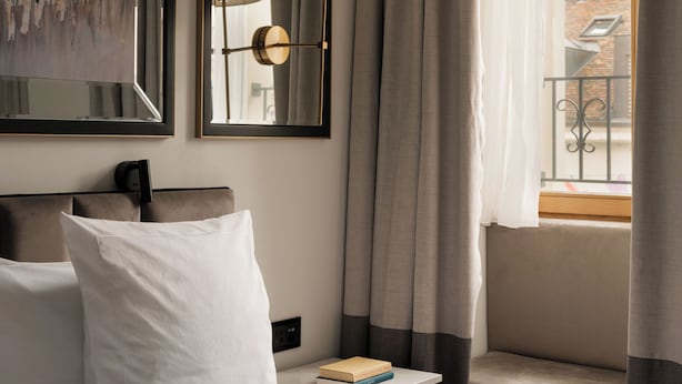 Una habitación de hotel con una cama, una mesita de noche con un libro, una lámpara, un espejo grande y una ventana con cortinas.
