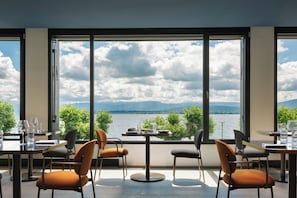 Comedor con una mesa dispuesta para cuatro, ventanas amplias con vista a un lago y montañas, y sillas con cojines.