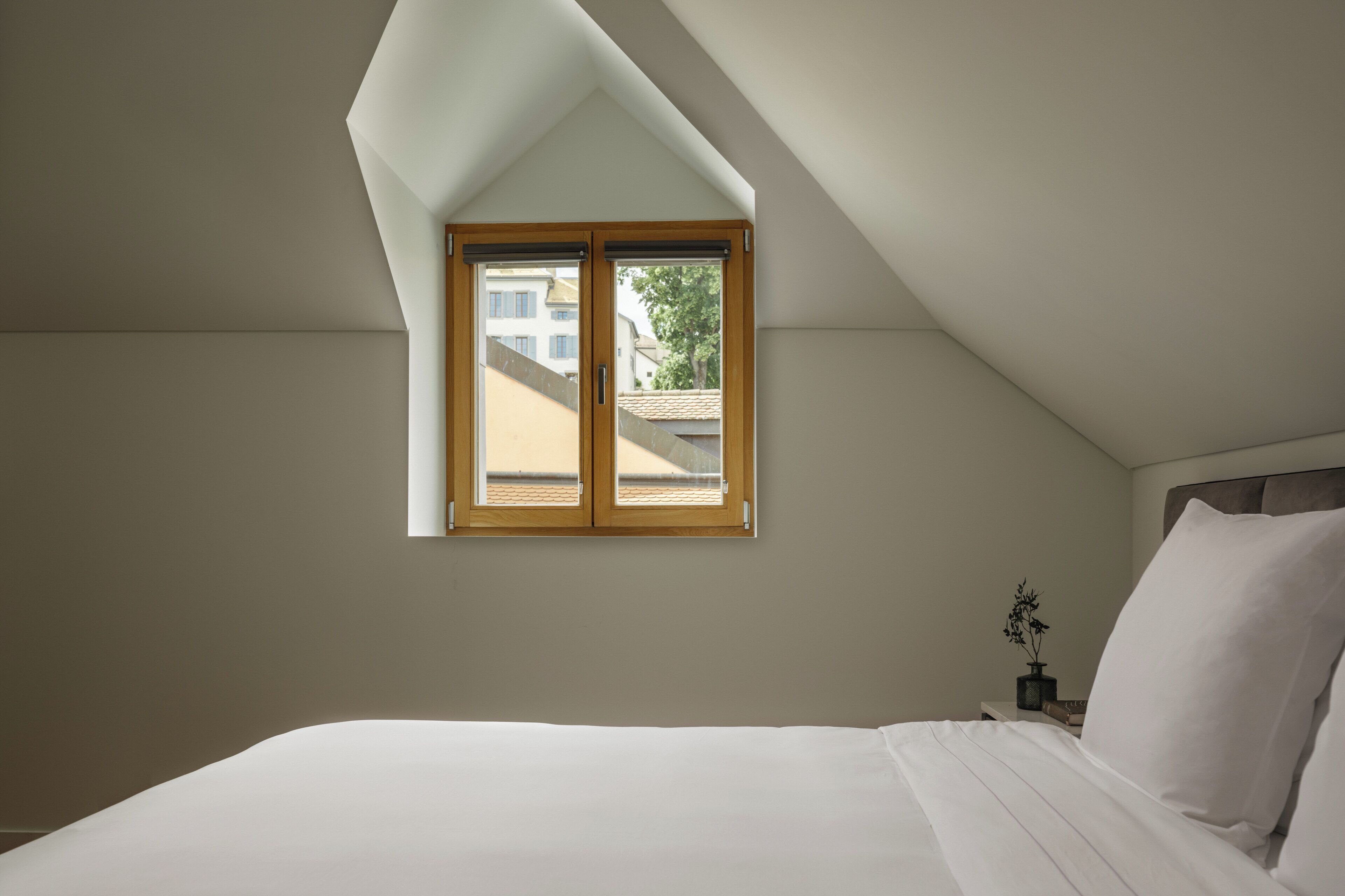 Une chambre avec un plafond en pente, une fenêtre, un lit, une table de chevet et une plante en pot.