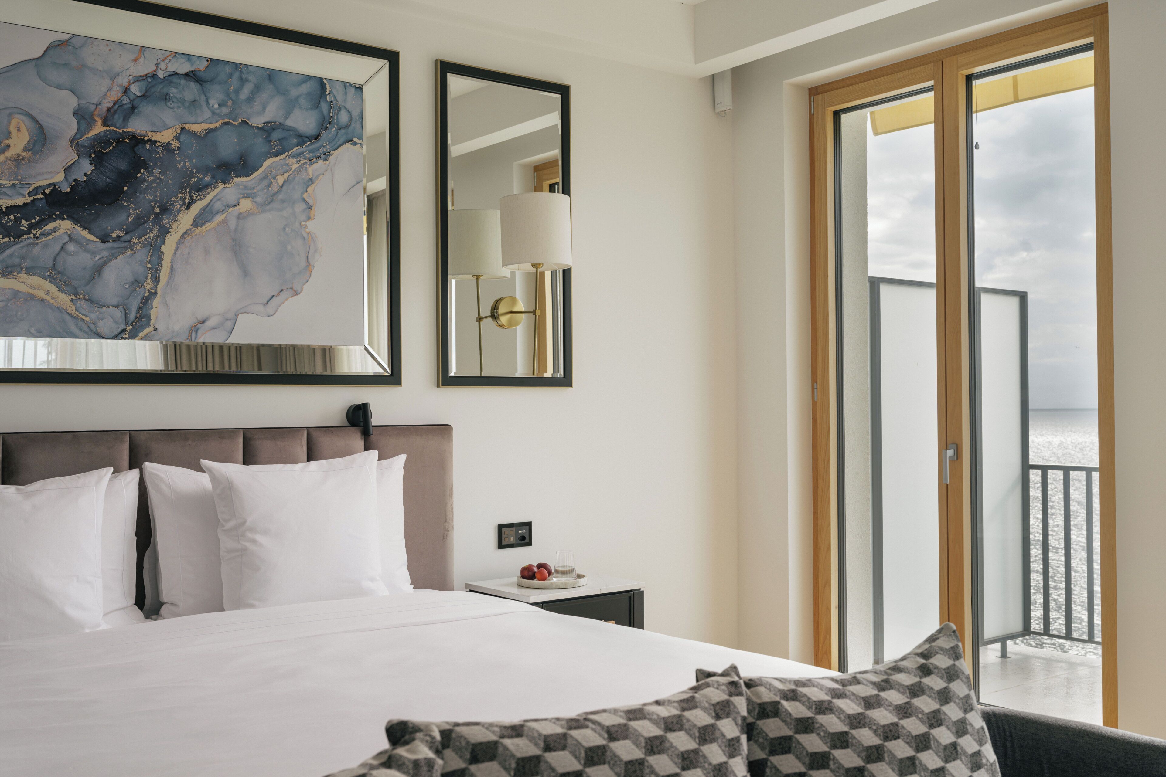 A hotel room with a large bed, a nightstand with a lamp, a framed artwork, and a view through a sliding door.