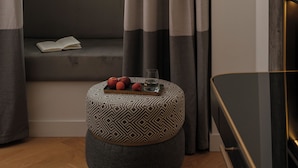 A room with a window, curtains, a sofa, a small ottoman with apples and a glass, and a wall-mounted light fixture. - Beau-Rivage Nyon (Nyon)