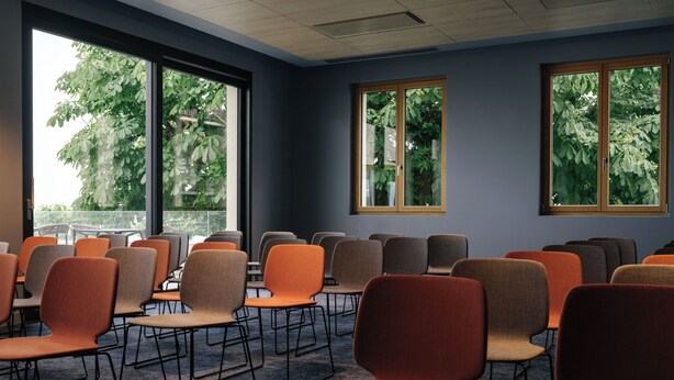 A conference room with rows of chairs and tables, large windows with views of trees, and a ceiling with recessed lighting. - Beau-Rivage Nyon (Nyon)