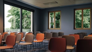 Un salón de conferencias con filas de sillas y mesas, ventanas amplias que dan a vistas de árboles e iluminación empotrada en el techo.
