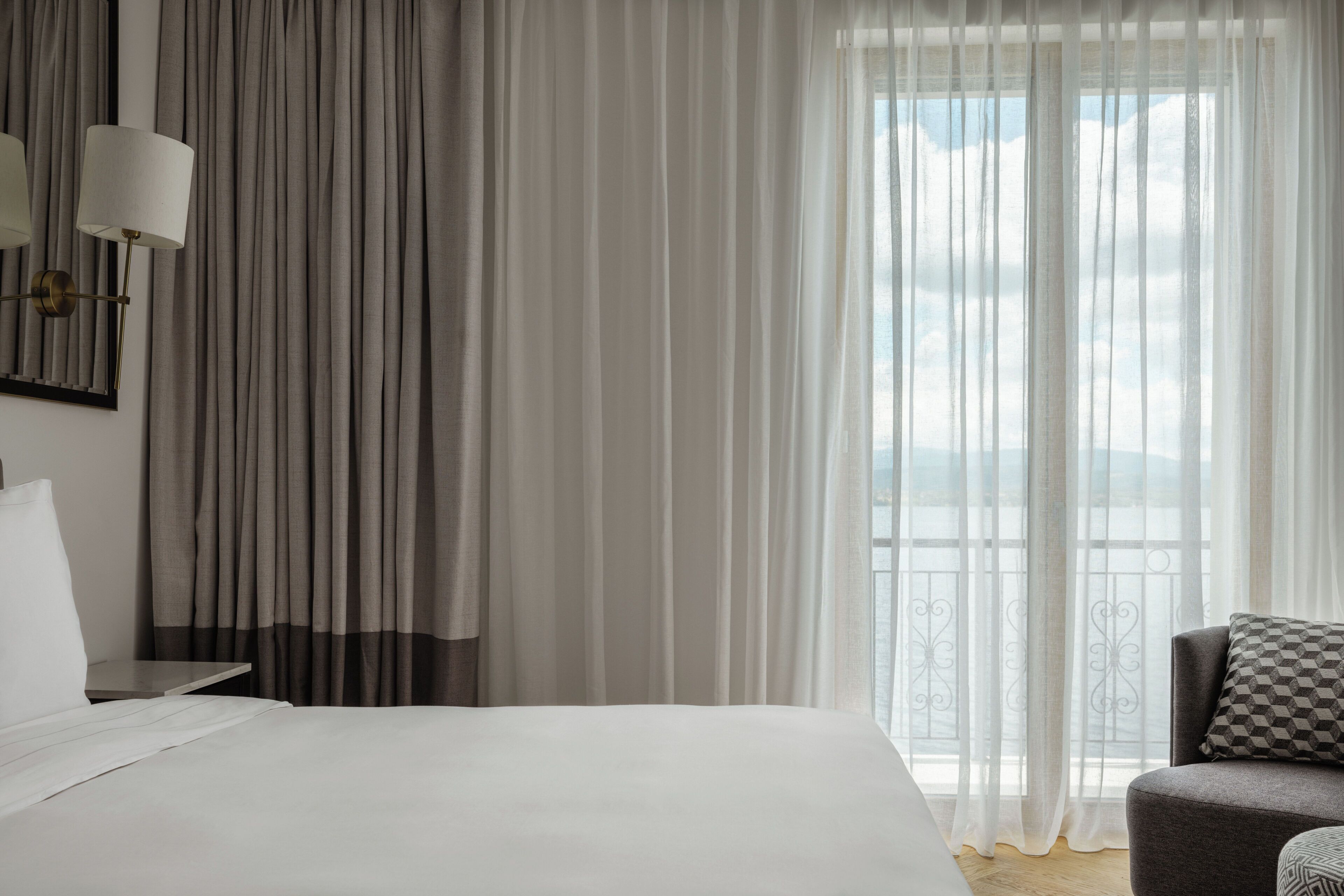 A hotel room with a bed, a chair, a nightstand, and sheer curtains.