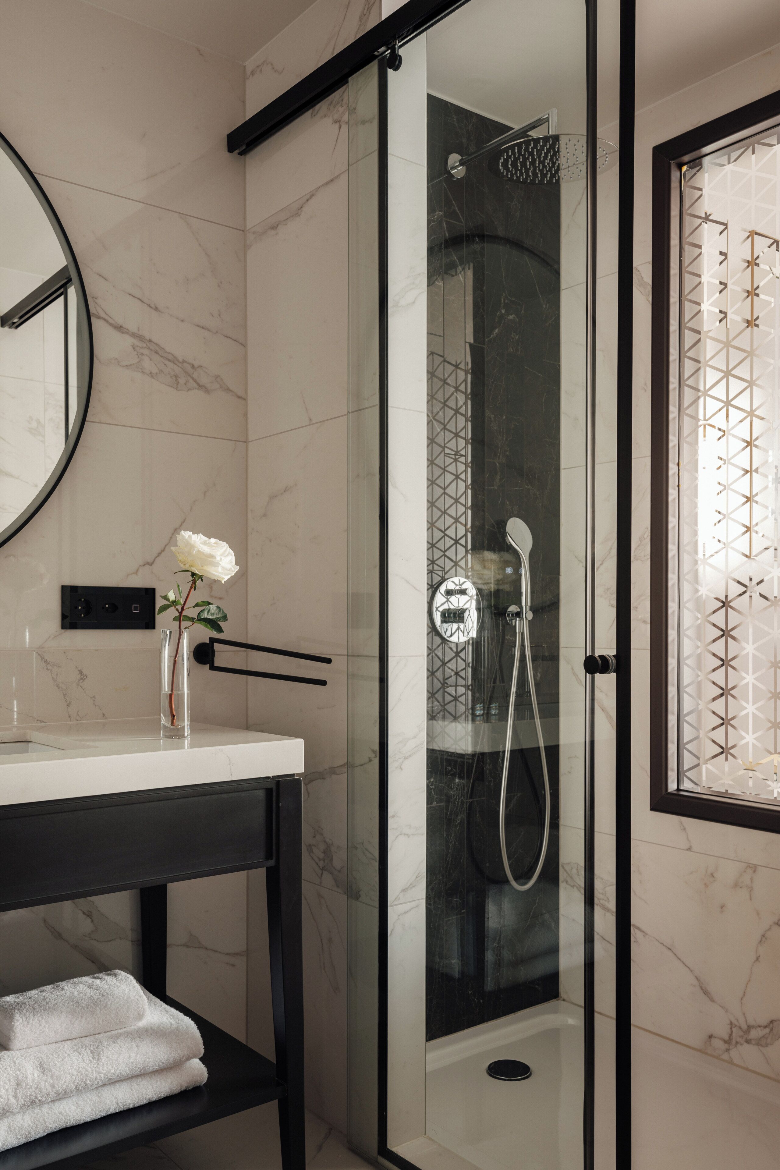 Une salle de bain moderne avec une douche à parois de verre, un mur recouvert de carreaux de marbre, un miroir rond et un vase contenant une seule rose blanche.